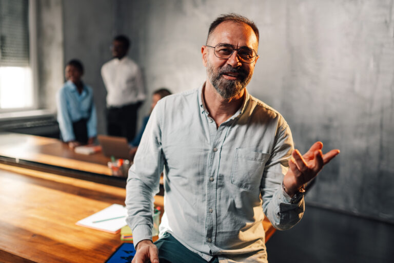 Friendly mature manager talking to camera and gesturing at boardroom